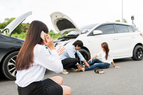 woman on the phone while the other driver and passenger are on the ground with injuries. Black car and white car head on collision